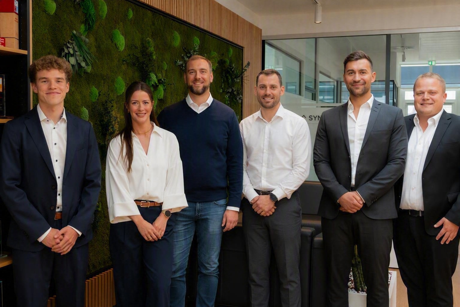 Bild mit 6 Personen vor einem Hintergrund im Büro, lächeln in die Kamera.
